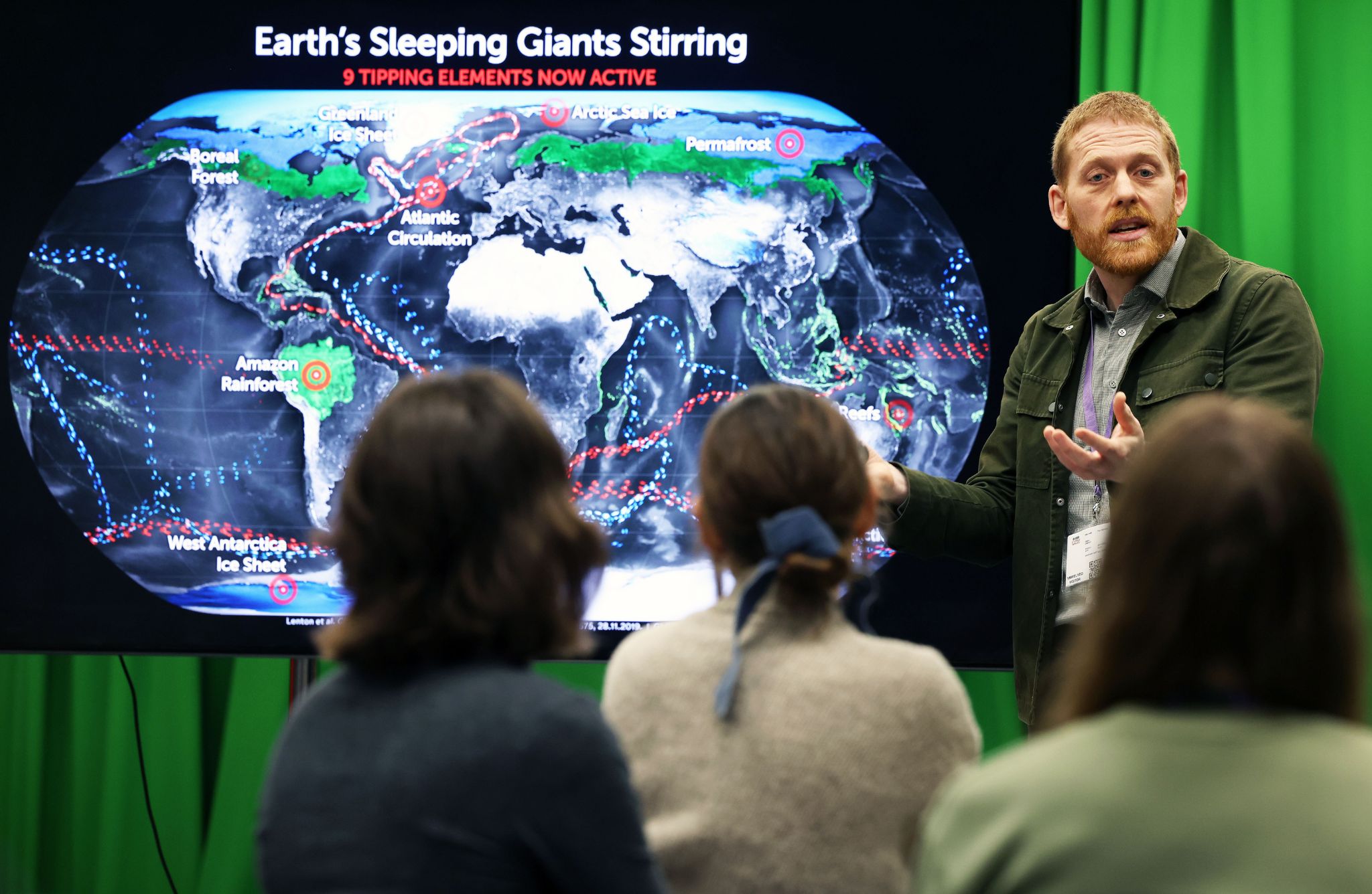 A person is giving a presentation in front of a large screen displaying a world map titled “Earth’s Sleeping Giants Stirring” with the subtitle “9 Tipping Elements Now Active.” The map highlights regions such as the Amazon rainforest, Atlantic circulation, West Antarctic ice sheet, and permafrost, with colored markers and dashed lines indicating climate-related tipping points. Three individuals are seated in the foreground, facing the presenter. The background includes a green curtain.