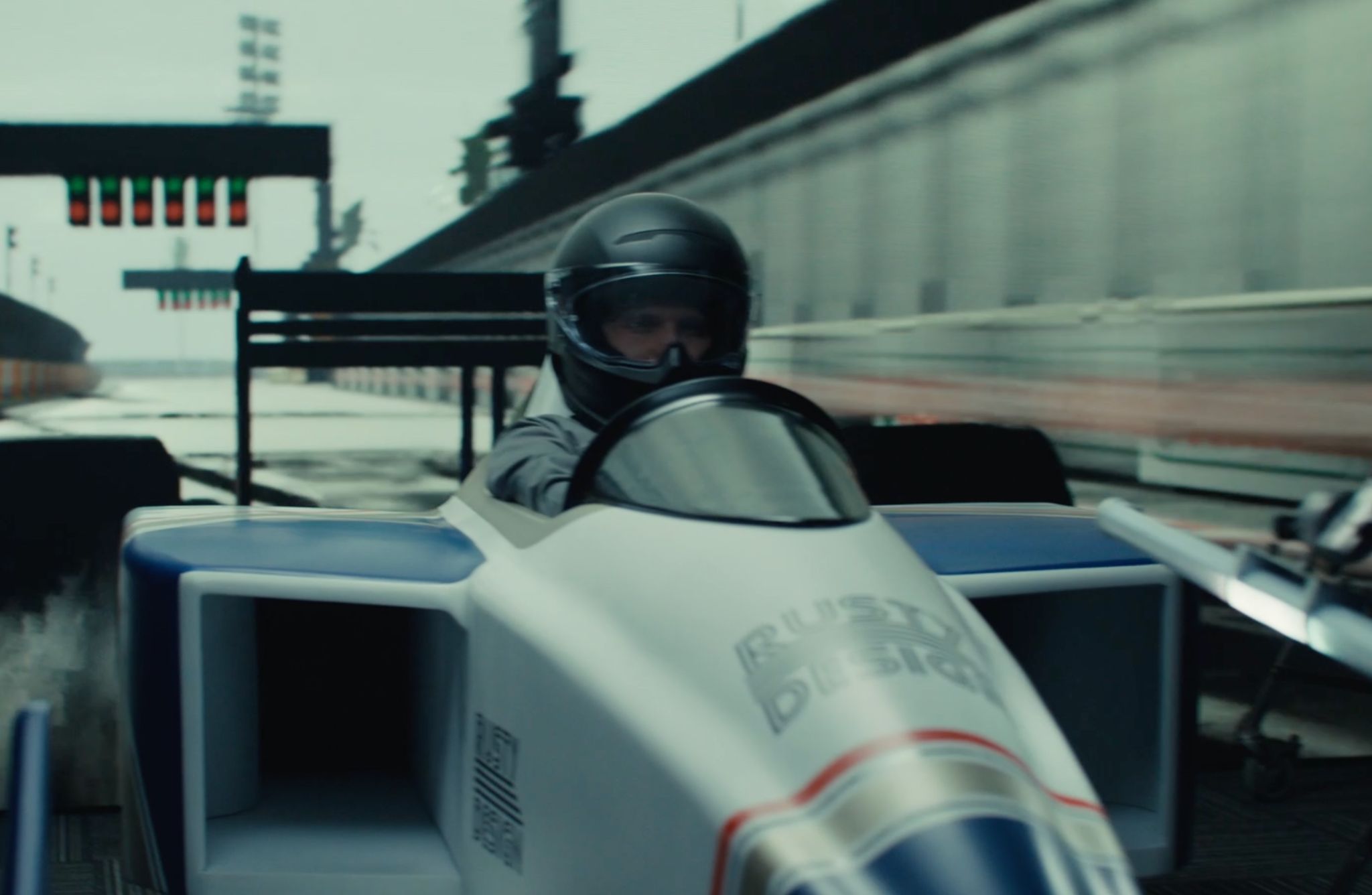 A sleek, single-seat race car with a white and blue body is positioned on a racetrack. The car features aerodynamic side panels and visible branding on the front. The driver is wearing a black helmet and racing suit. In the background, there is a blurred view of the pit lane and a starting light structure with illuminated signals.