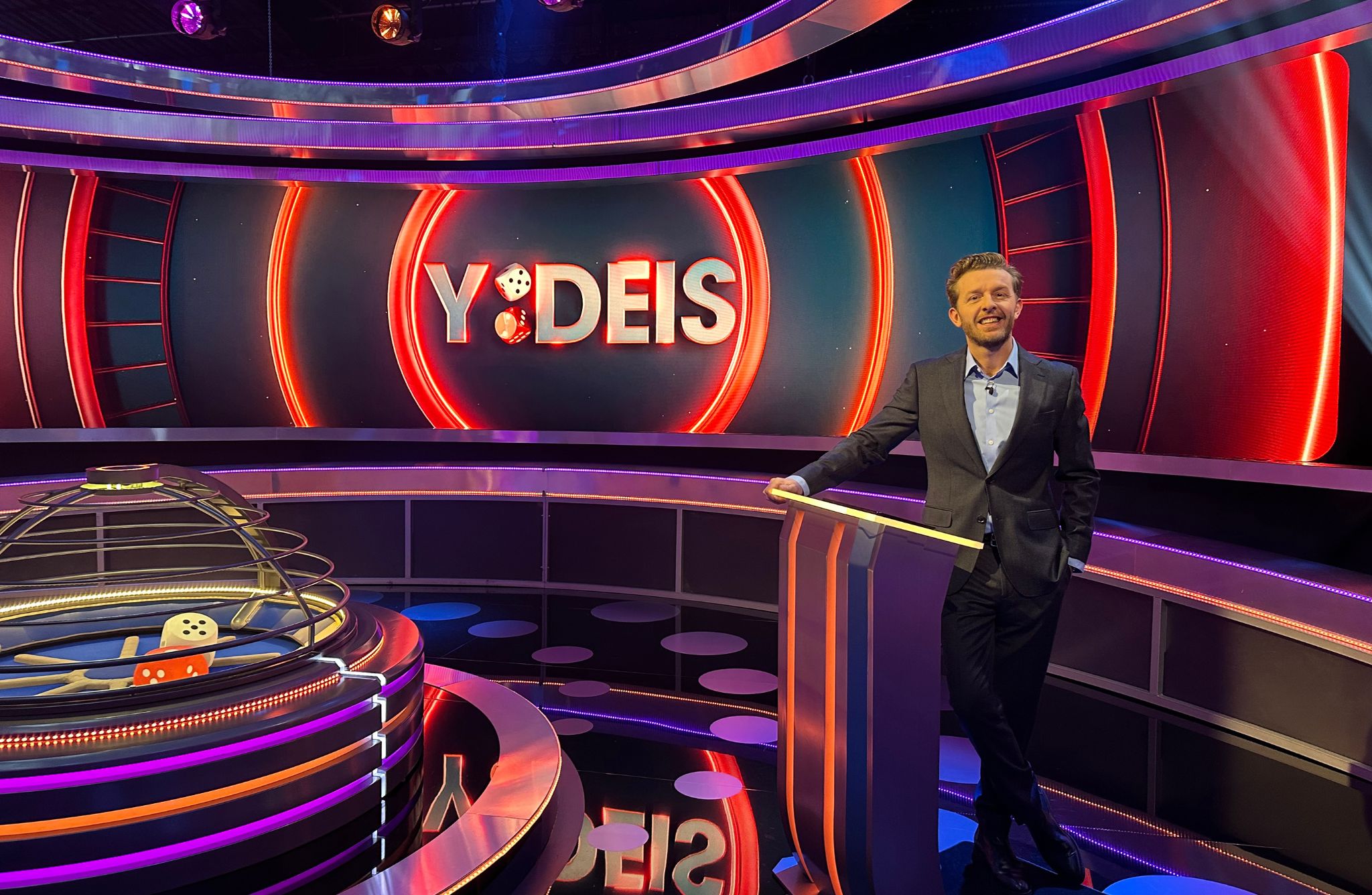 Game show set with neon lights, a podium with a person standing by it and smiling, and a large screen displaying text ‘Y DEIS’.
