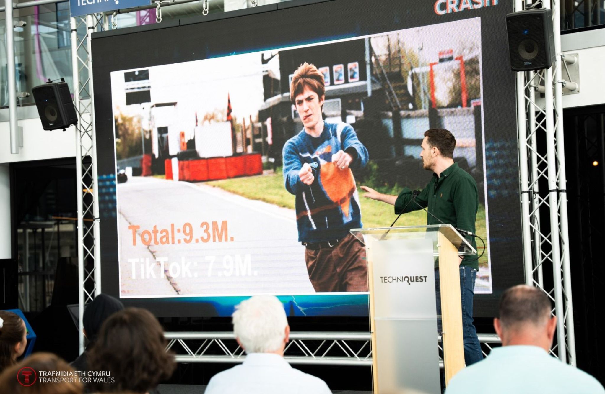 A person stands at a podium with a microphone, pointing toward a large screen behind them. The screen displays an image of someone holding an orange item outdoors, with text overlaid that reads “Total: 9.3M” and “TikTok: 7.9M.”