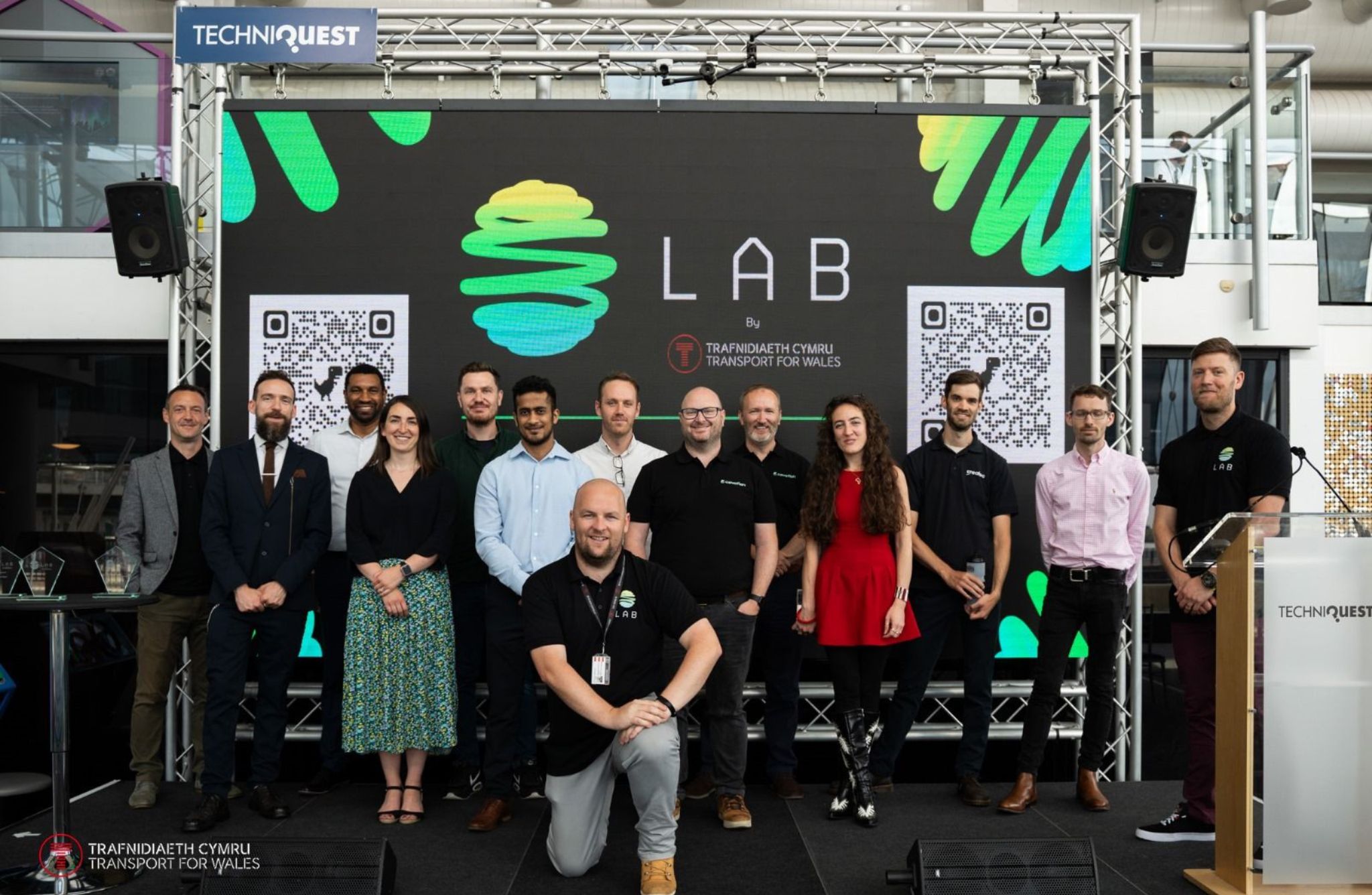 A group of people standing and kneeling on a stage in front of a large digital screen displaying the word “LAB” with a stylized green and yellow abstract logo.