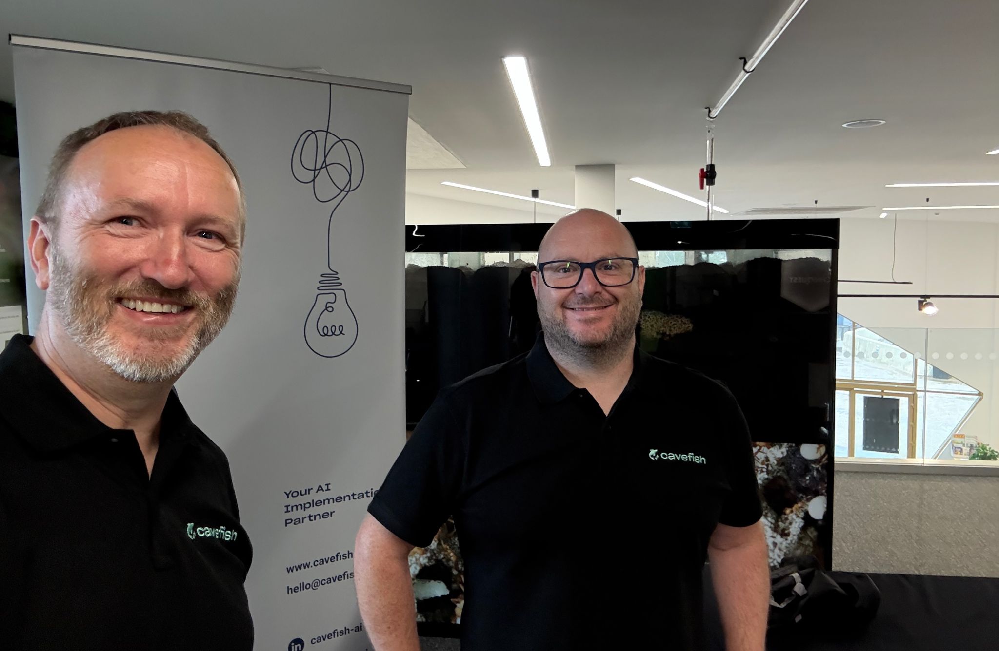 Two individuals wearing black polo shirts with the “Cavefish” logo stand indoors near a display booth. Behind them is a banner featuring a minimalist lightbulb illustration with a tangled cord and the text “Your AI Implementation Partner,” along with contact details and the Cavefish website.