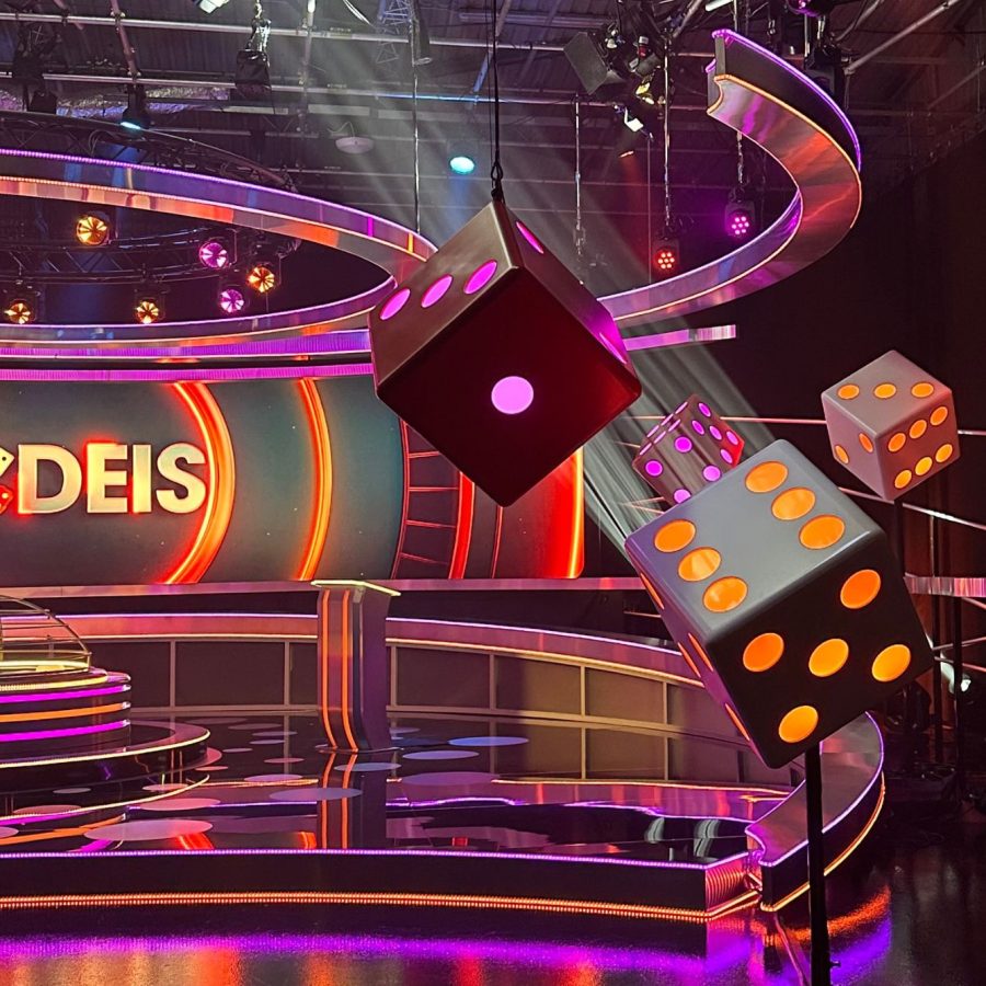 Colorful game show set with neon lights, large dice decorations, and “Y DEIS” displayed on the backdrop.