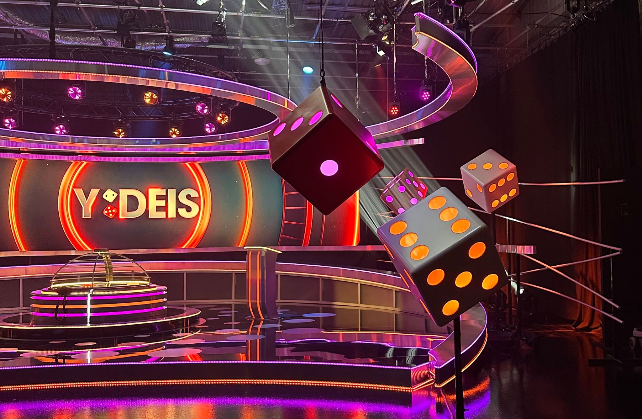Colorful game show set with neon lights, large dice decorations, and “Y DEIS” displayed on the backdrop.