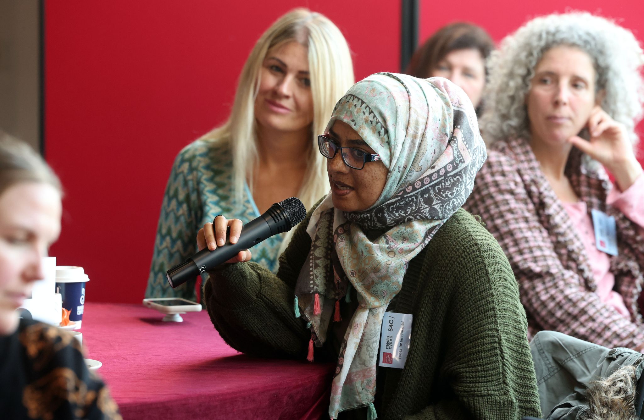Several people sit around a table covered with a red cloth during a discussion. One person in the foreground wears a patterned headscarf and green sweater, holding a microphone. A coffee cup and small dish are visible on the table, with a bright red background behind the group.