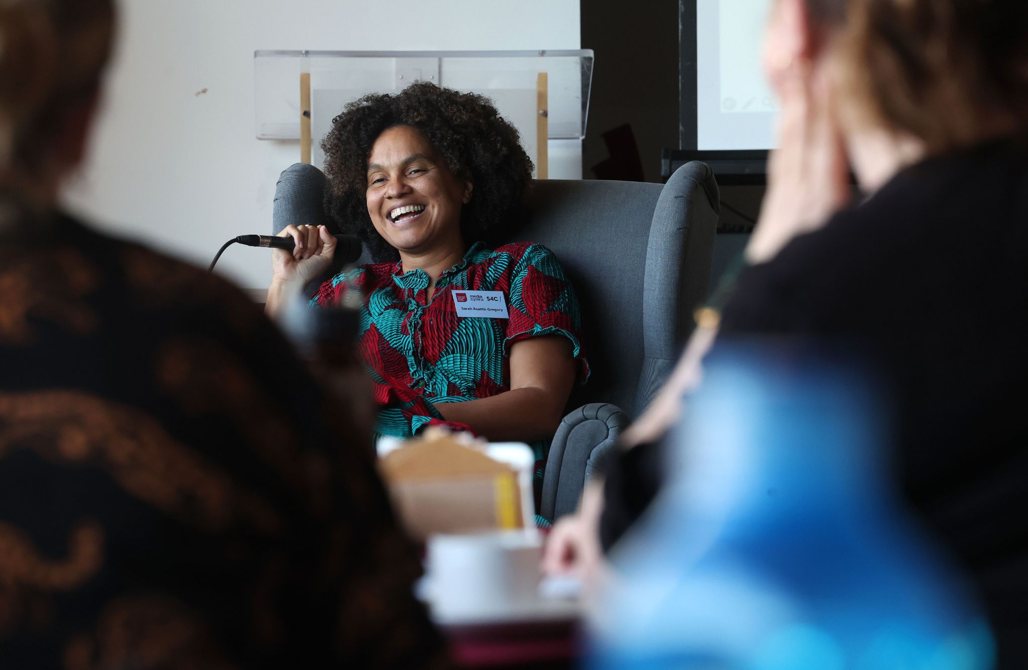 A person sits in a gray armchair holding a microphone during a discussion. The individual wears a red and green patterned top. In the foreground, blurred figures and a table with cups and a water bottle are visible, suggesting a group setting or event.