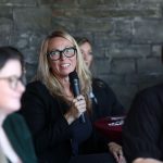 Person in black clothing holds a microphone while seated at a table with others. A stone wall and red tablecloth are visible in the background.