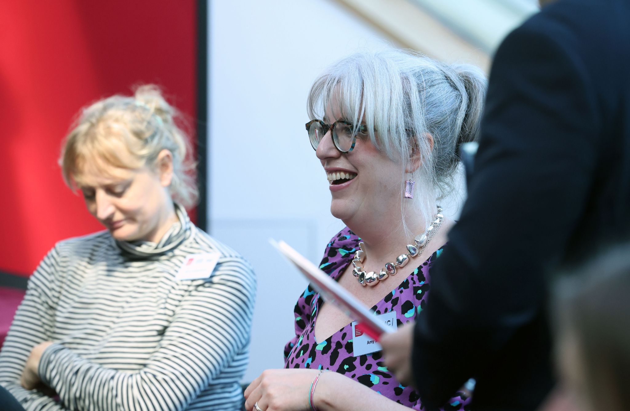 Two people seated indoors, one in a striped top and the other in a purple patterned outfit with a chunky necklace, while a third person stands holding a document.