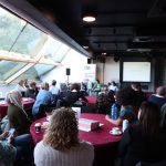Audience seated at round tables listens to a panel discussion in a modern venue with large windows, Media Cymru banner, and projection screen visible.