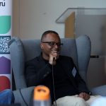 Person seated in a gray armchair speaks into a microphone during a discussion. A Media Cymru banner and a podium are visible in the background.