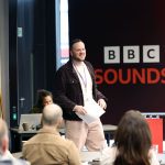 A speaker stands at the front of a room holding papers while addressing an audience seated at tables. A large BBC Sounds banner is displayed behind them