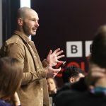 A person standing near a large screen speaks to a seated audience. The background shows BBC branding.