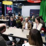 A larger group of people seated around multiple tables in a modern studio space with BBC branding visible. Individuals appear engaged in conversation and discussion.