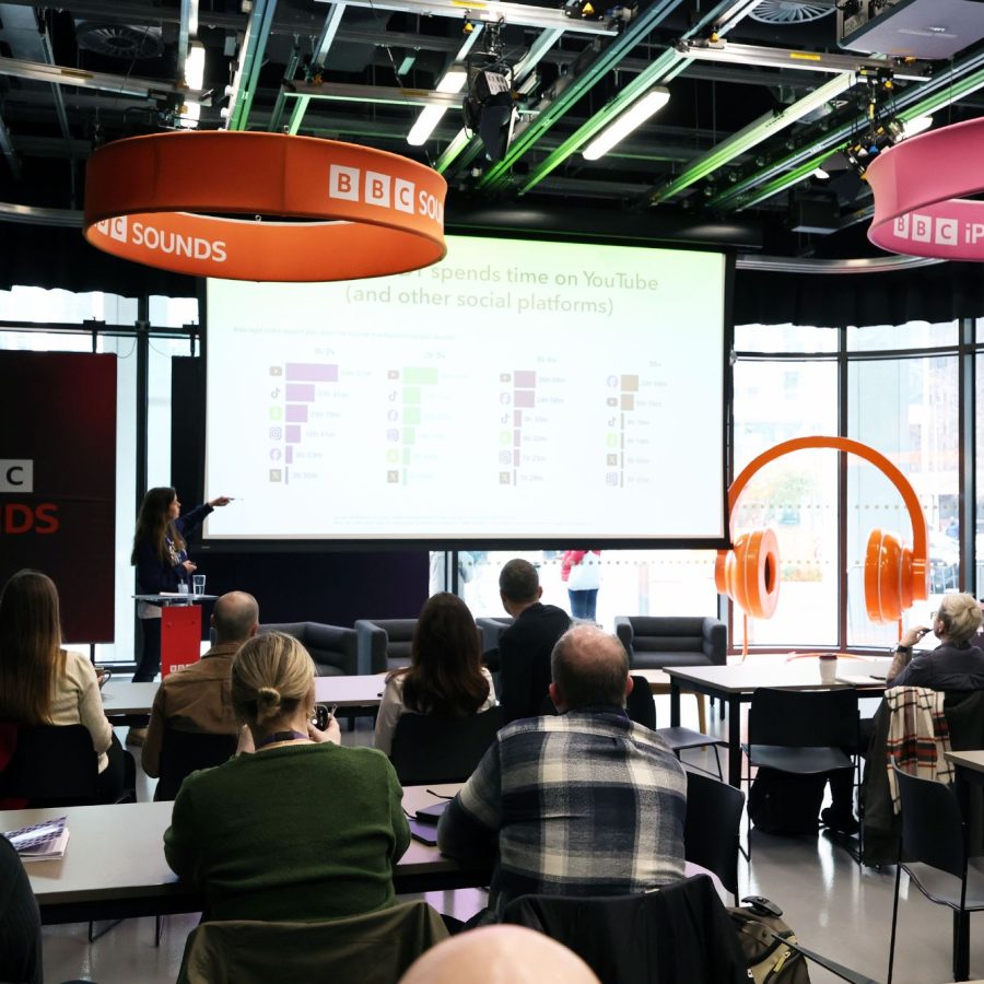 People sit in a BBC event space watching a presenter speak in front of a screen displaying charts.