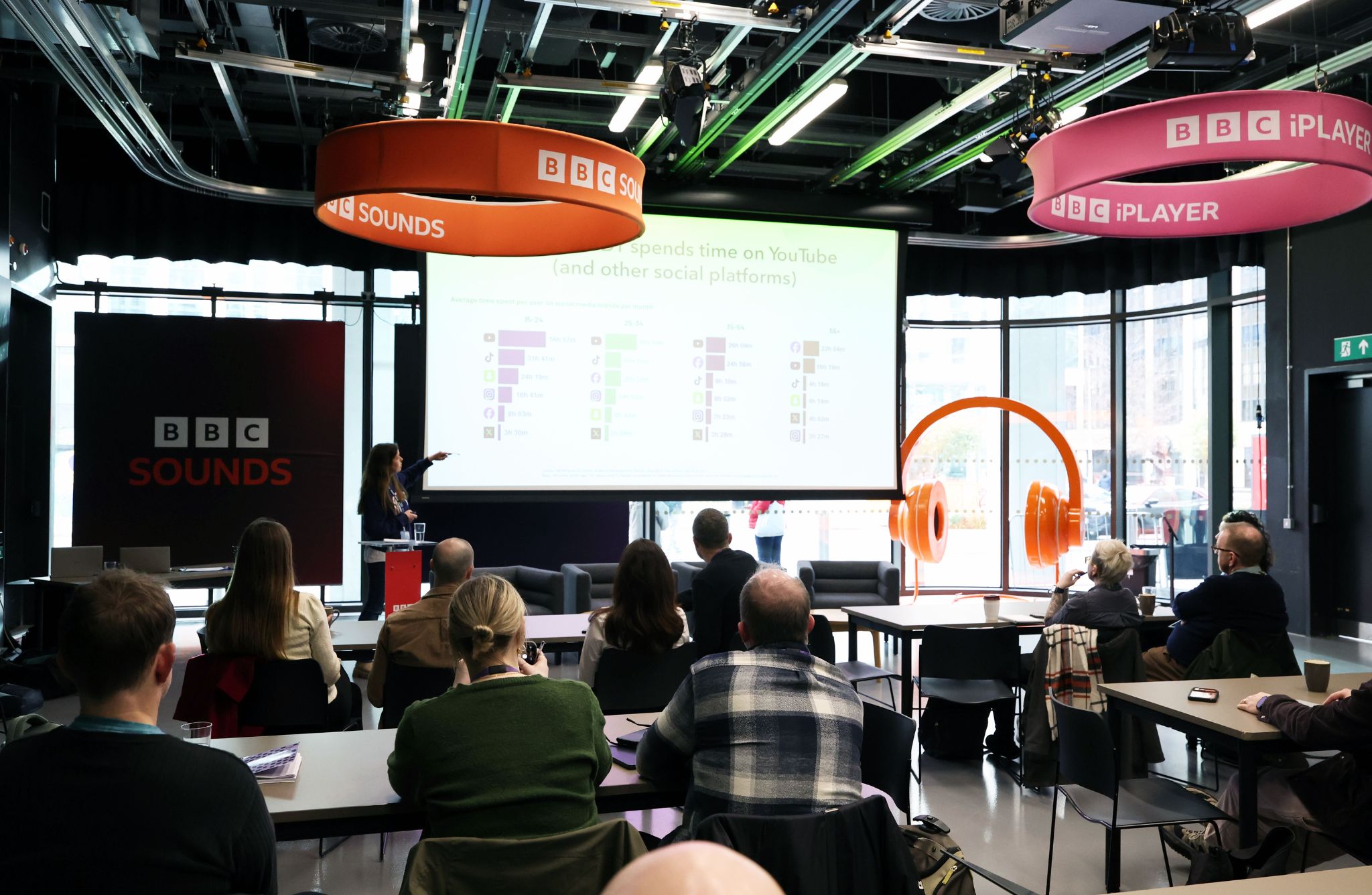 People sit in a BBC event space watching a presenter speak in front of a screen displaying charts.