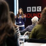 A speaker stands at the front of a room giving a presentation, with attendees seated facing them. A large BBC Sounds backdrop is visible behind the speaker.