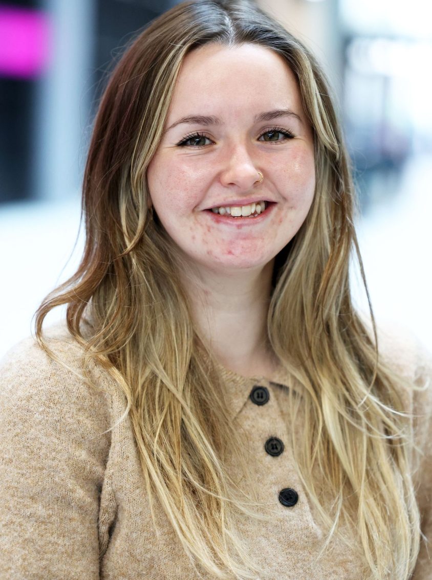 A person with long, wavy, light brown and blonde hair wearing a tan knit sweater with three dark buttons. They are standing indoors in a softly lit space with a blurred background.