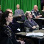 A classroom-style setup where people sit at desks facing forward. Open notebooks, mugs, and lanyards are visible, with a green curtain in the background.