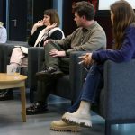 A small panel of four people seated in armchairs on a stage or discussion area. One person speaks while the others listen.