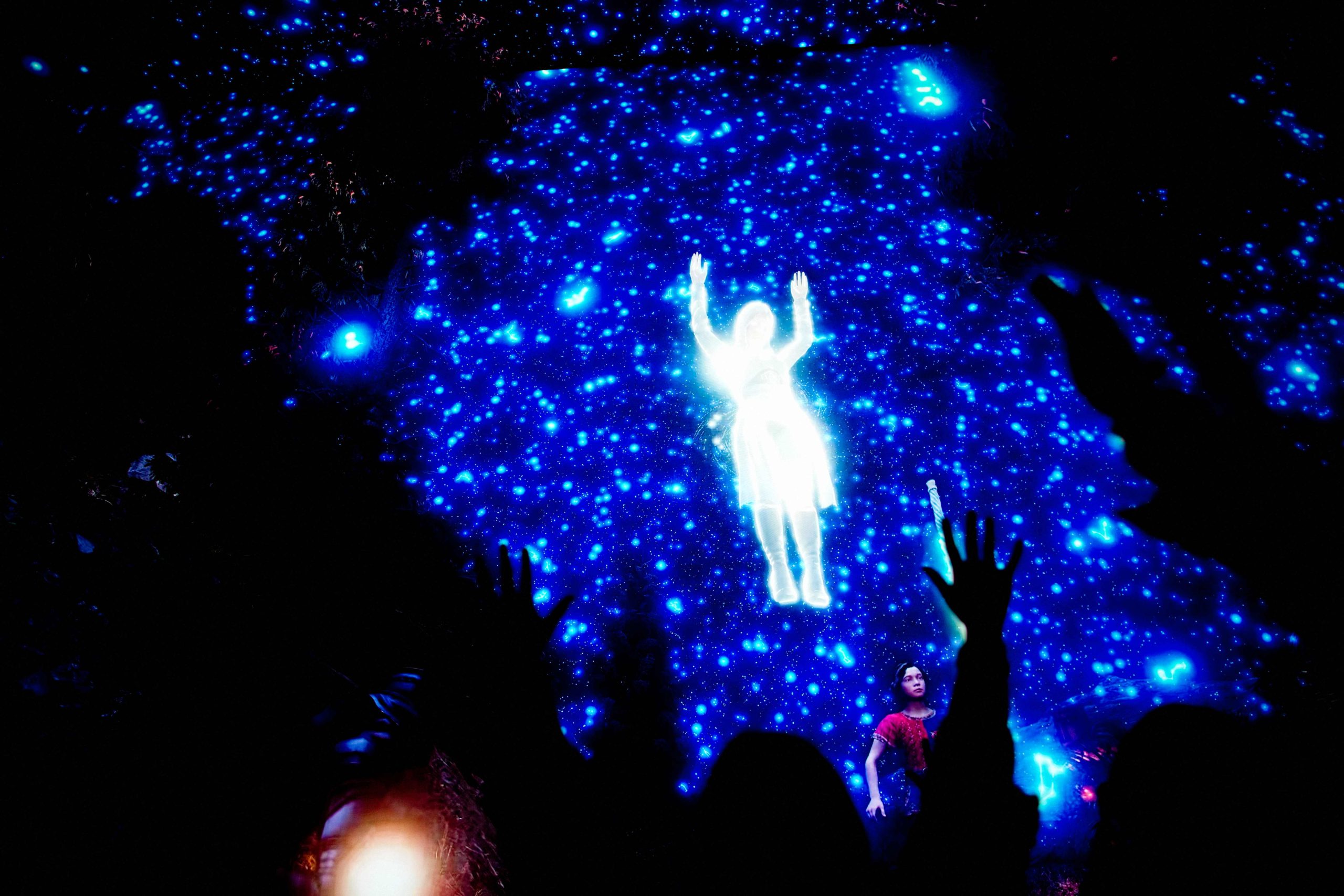 A group of silhouetted people stand with raised arms facing a large, immersive digital display. The screen shows a bright, glowing figure suspended against a backdrop of deep blue light filled with star‑like points. One person stands closer to the screen, illuminated by the projection, while the rest of the crowd is mostly in shadow. The scene has a dramatic, otherworldly atmosphere created by the intense contrast between darkness and the radiant figure.