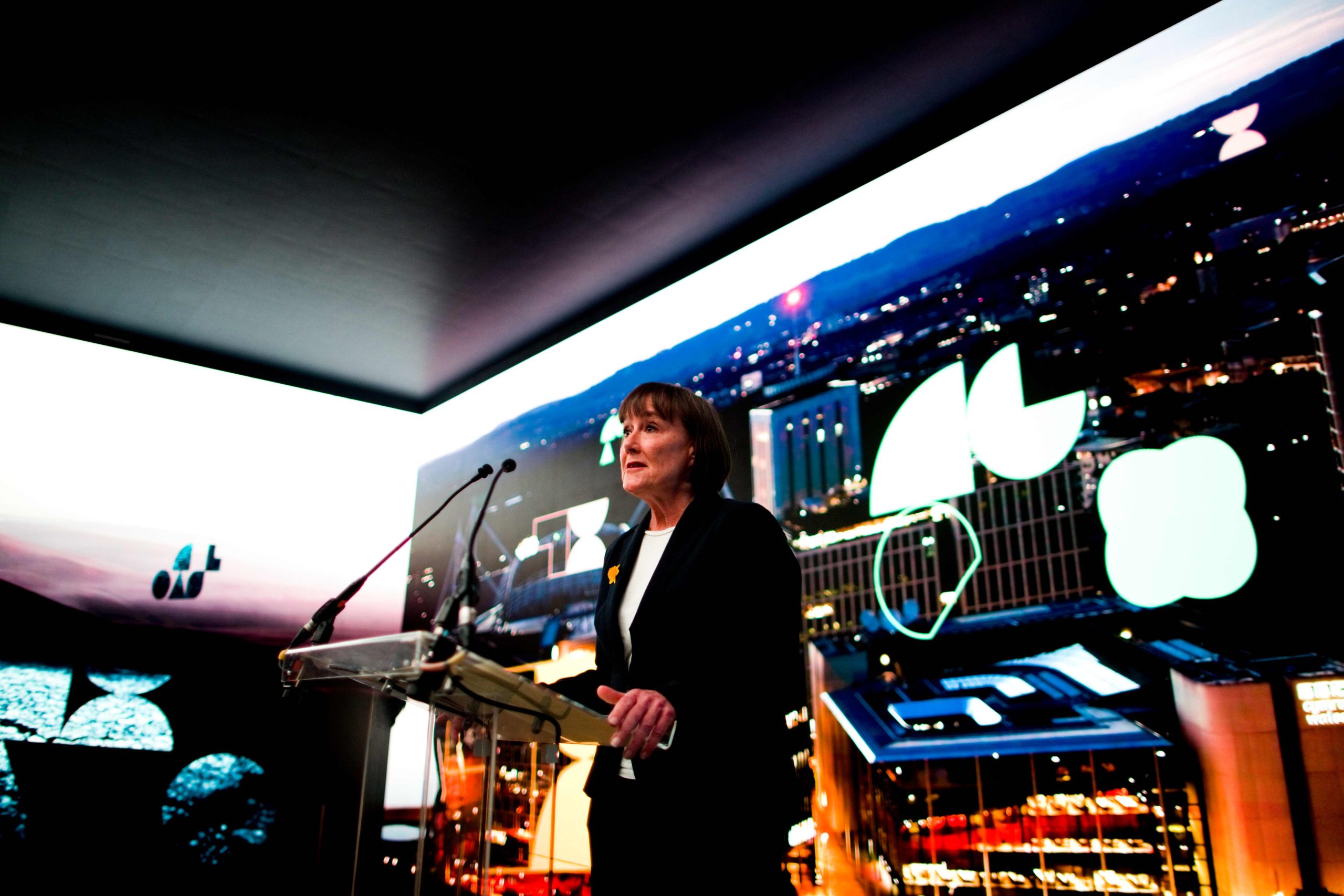 Jo Stevens MP stands at a transparent podium with two microphones, delivering a speech in front of a large wraparound digital screen. The screen displays a nighttime cityscape with illuminated buildings, mountains in the distance, and several large abstract graphic shapes layered over the image. The lighting in the space is low, with the digital backdrop brightly illuminating the speaker from behind.
