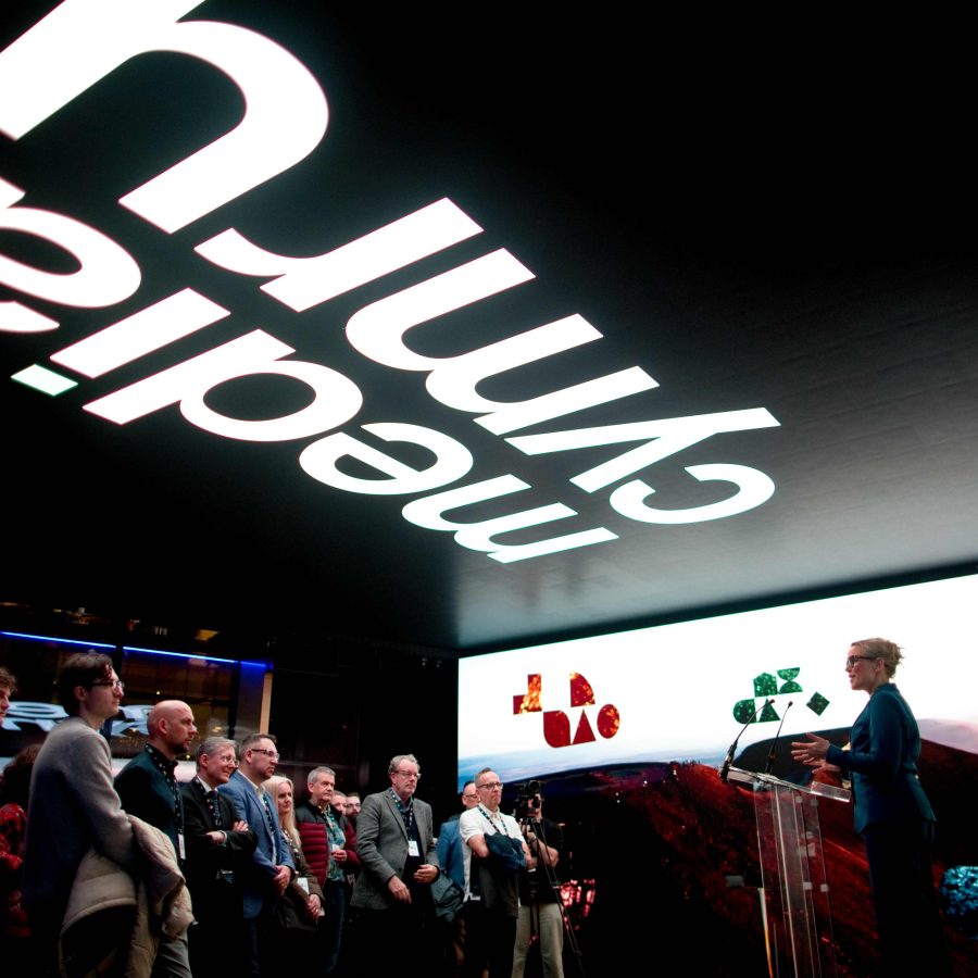 A person stands at a clear podium on a stage, speaking to an audience gathered in a dimly lit event space. A large illuminated screen behind the speaker displays abstract colorful shapes over a landscape image. Above the crowd, a ceiling installation features large, inverted white text reading “Media Cymru.” Members of the audience stand in a semi-circle facing the stage.