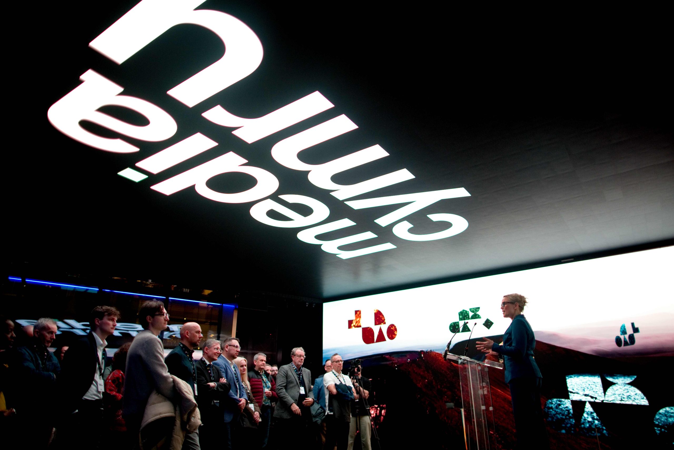 A person stands at a clear podium on a stage, speaking to an audience gathered in a dimly lit event space. A large illuminated screen behind the speaker displays abstract colorful shapes over a landscape image. Above the crowd, a ceiling installation features large, inverted white text reading “Media Cymru.” Members of the audience stand in a semi-circle facing the stage.