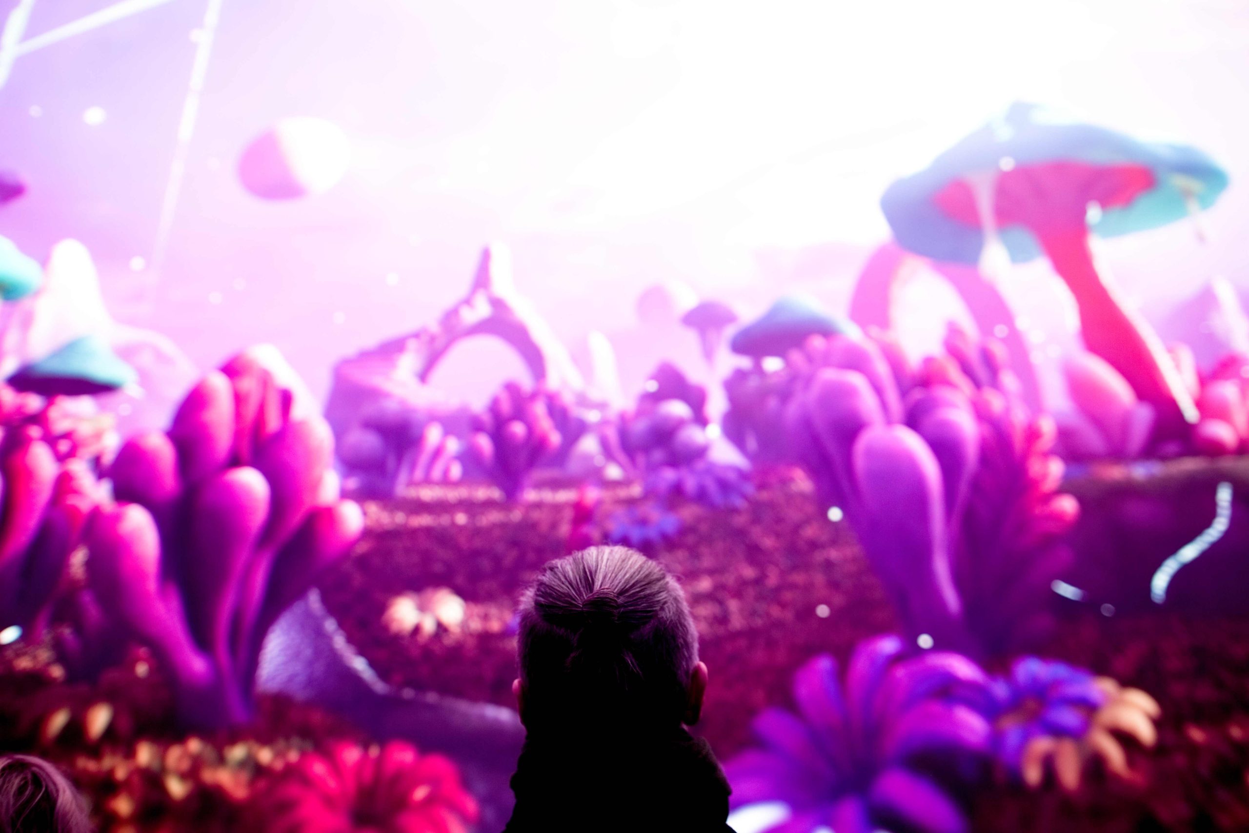 A person stands in the foreground facing a large, brightly lit screen that displays a vivid fantasy landscape. The scene features oversized, glowing mushrooms, bulbous pink and purple plants, and soft, surreal terrain under a bright, hazy sky.
