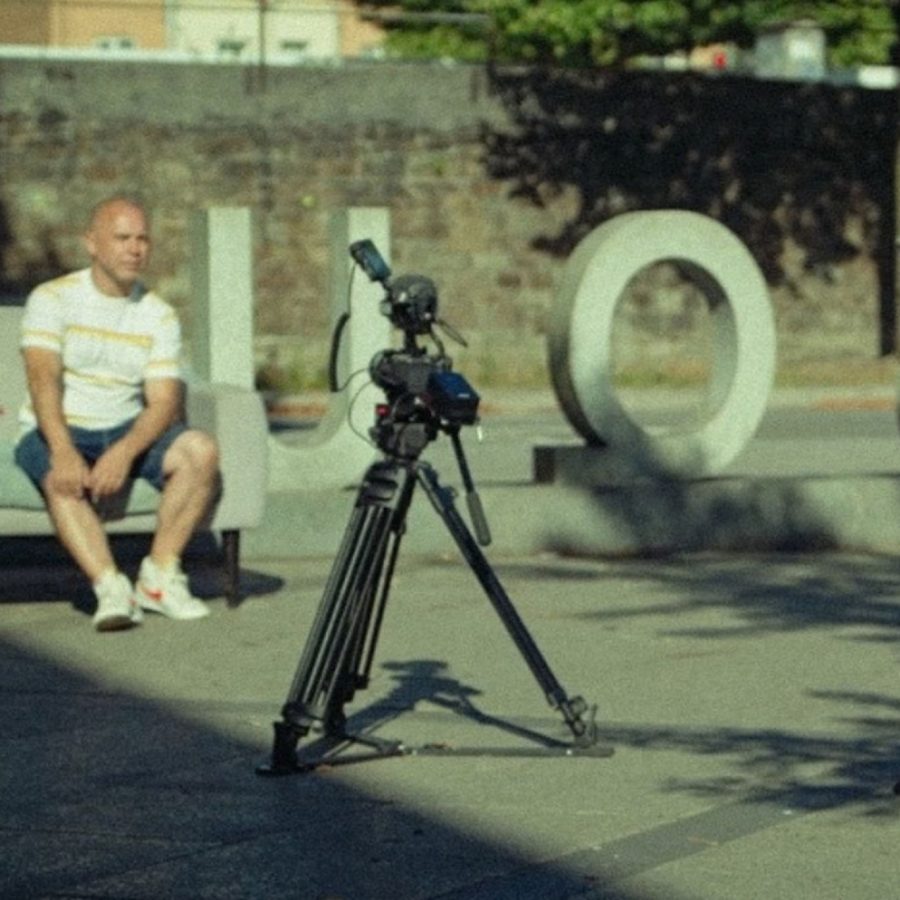 A person sits on an outdoor sofa in a shaded area while a professional video camera on a tripod is positioned in front of them. Large sculptural letters are arranged behind the scene, and trees and a stone wall are visible in the background.
