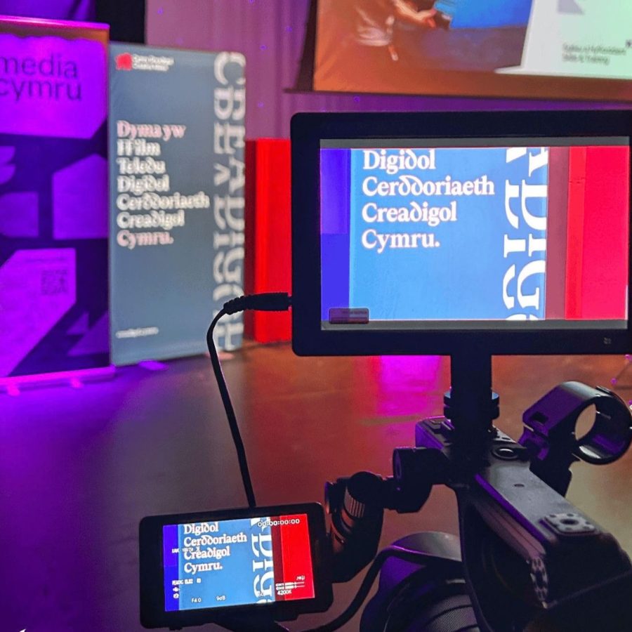 “A video camera records a stage setup with Media Cymru banners and a screen displaying Welsh text at an event.”