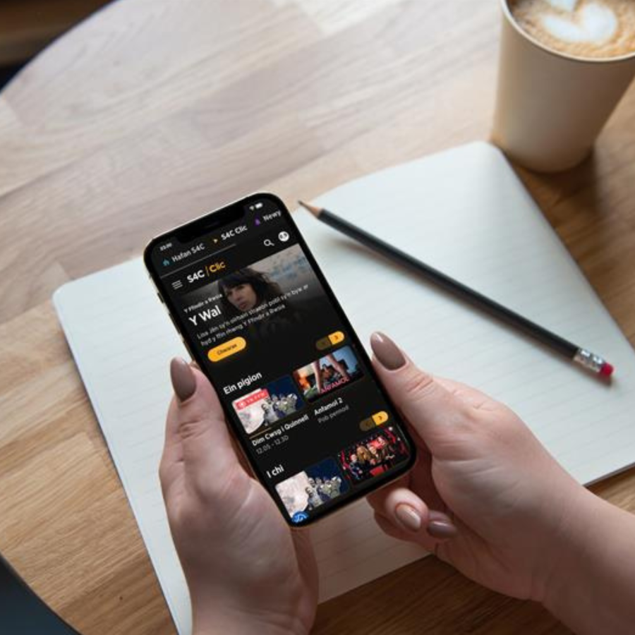 Hands holding a smartphone displaying a streaming app, resting above an open notebook on a wooden table with a pencil and a cup of coffee.