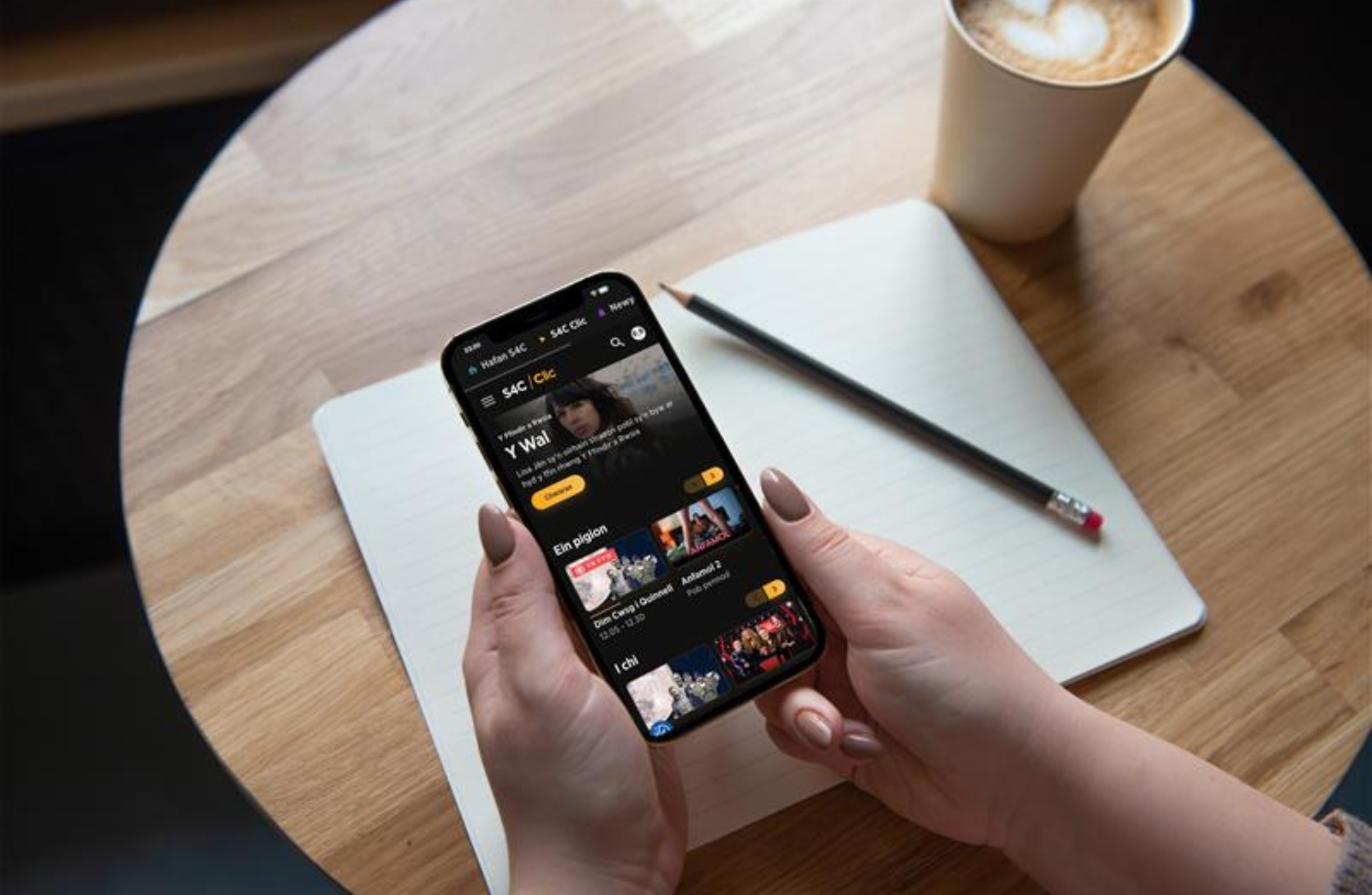 Hands holding a smartphone displaying a streaming app, resting above an open notebook on a wooden table with a pencil and a cup of coffee.