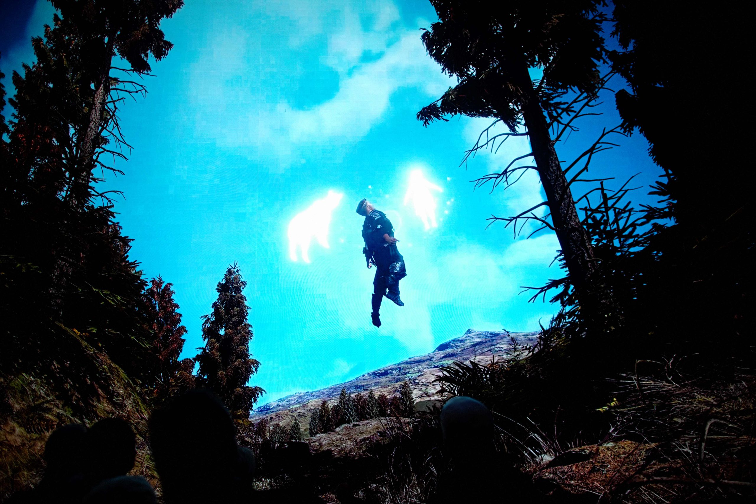 A figure appears to float above a forest clearing, framed by tall trees, with bright white animal‑like light forms glowing in a vivid blue sky behind them.
