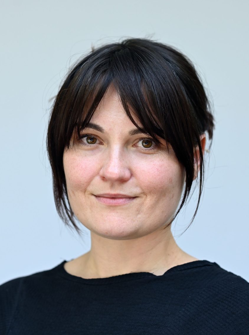 Head-and-shoulders portrait of a person with dark hair wearing a black top against a light grey background.